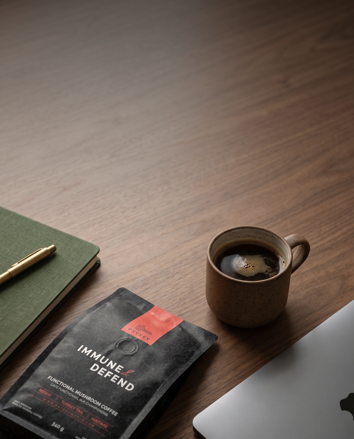 A bag of Immune Defend Functional Mushroom Coffee lies on a dark wood desk next to a brown ceramic mug of black coffee, a green notebook with a gold pen, and part of a silver laptop.