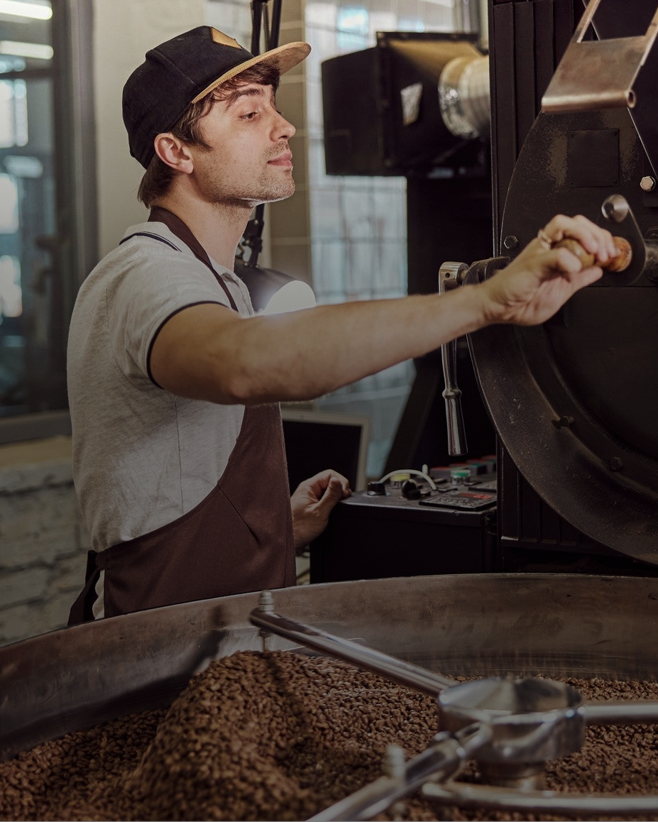 A roaster operator in a coffee roastery adjusts a lever on a large coffee roaster, with roasted coffee beans visible in the bin below.