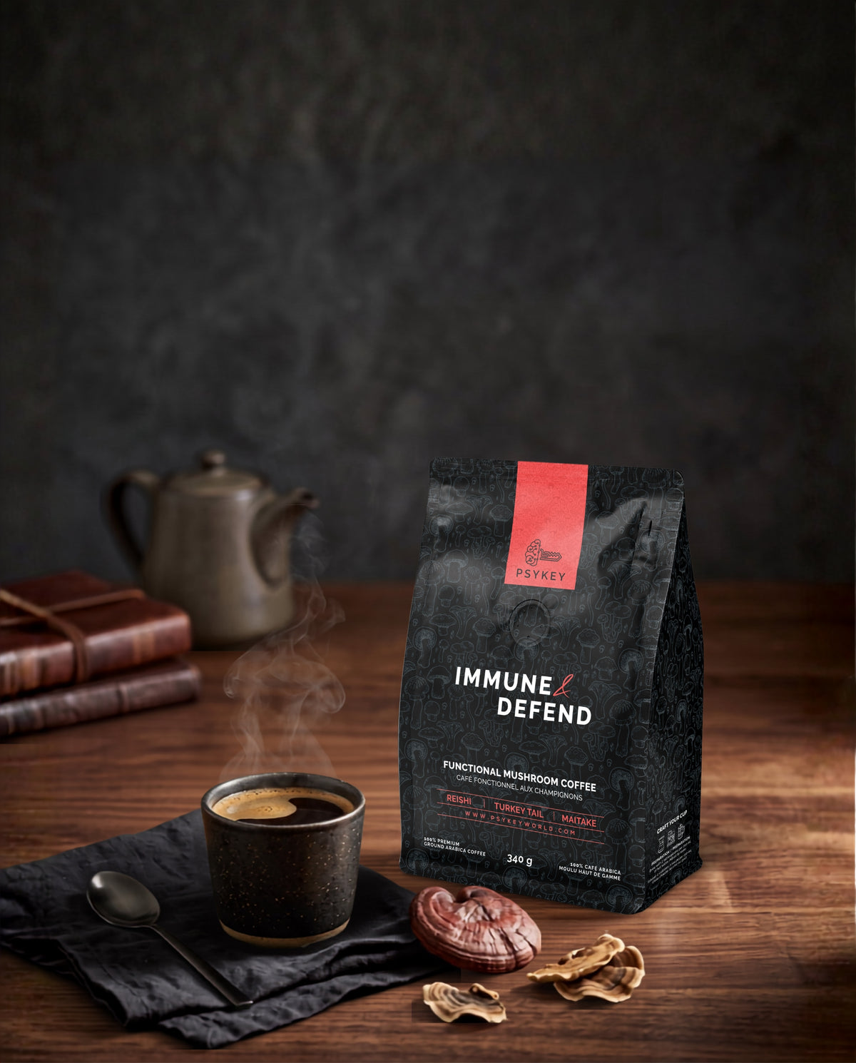 A bag of PsyKey brand "Immune & Defend" functional mushroom coffee sits on a rustic wooden table next to a steaming dark ceramic mug of black coffee, a small black spoon, and a stack of leather journals. A brown ceramic teapot rests softly in the blurred background. In the foreground, whole and sliced dried reishi mushrooms are scattered near the base of the sleek black coffee bag, which features a prominent red stripe at the top.