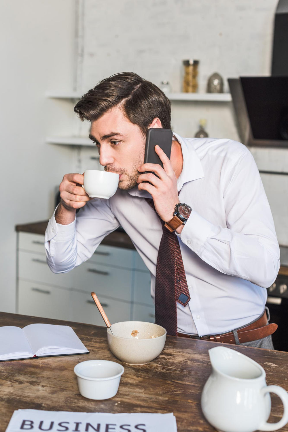 A professional man in a white dress shirt and tie leaning over a table, holding a phone to his ear while drinking a cup of coffee during his morning routine.
