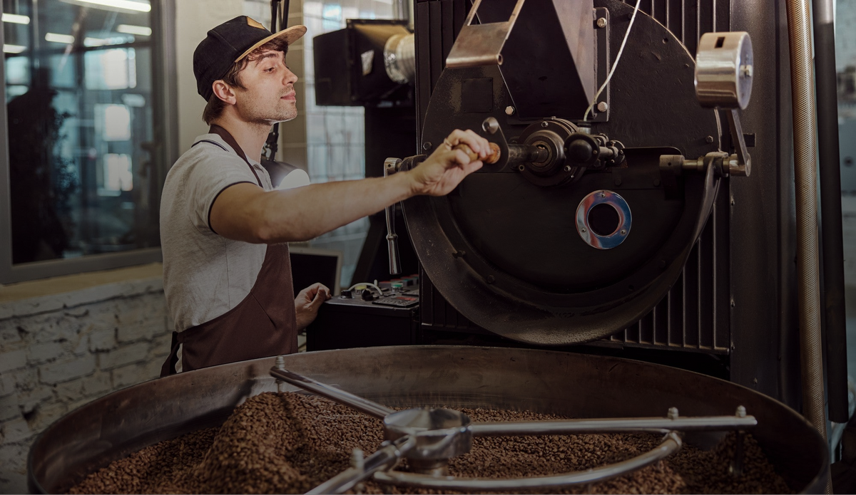 A roaster operator in a coffee roastery adjusts a lever on a large coffee roaster, with roasted coffee beans visible in the bin below.