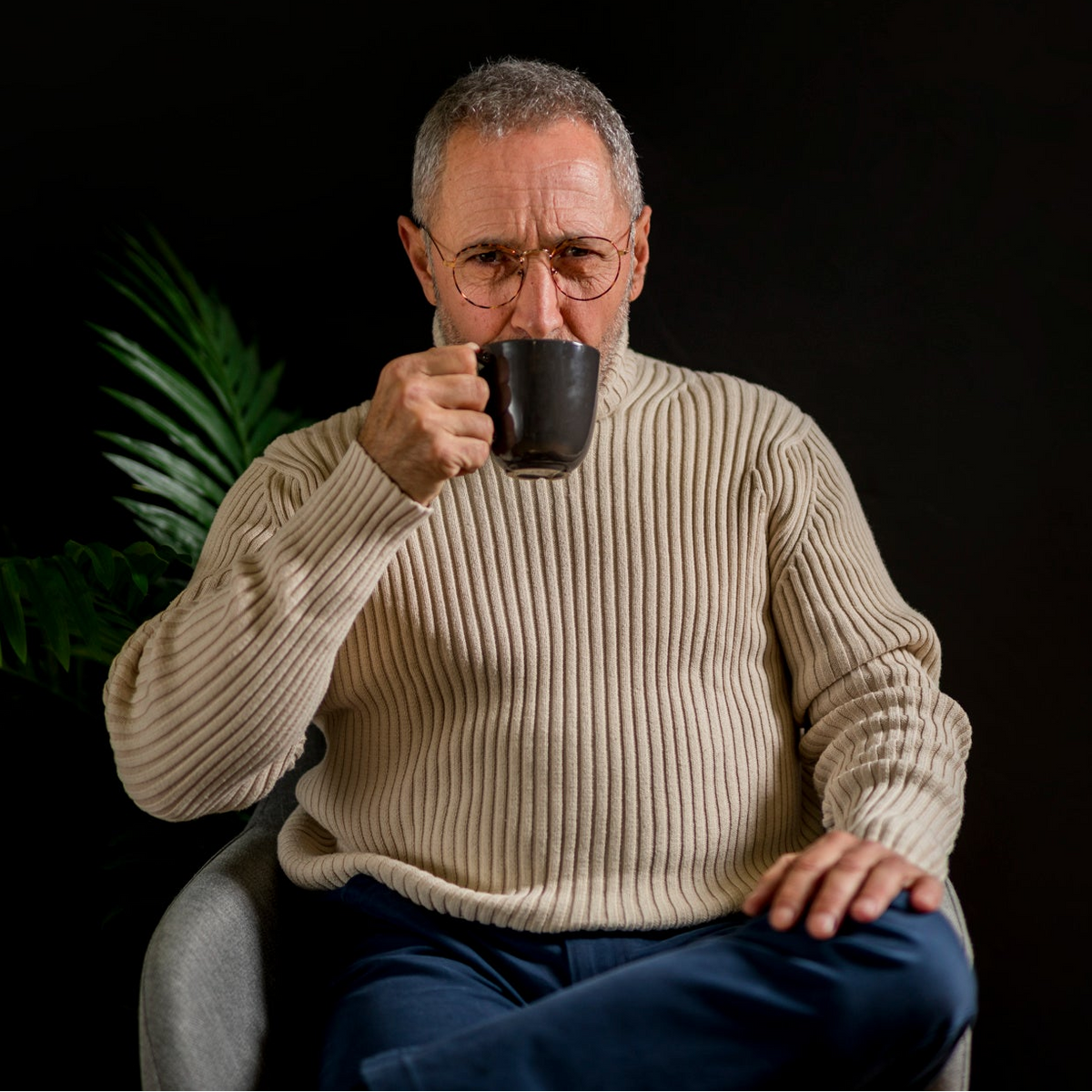 An older man with gray hair and glasses sits in a chair against a black background, holding a dark ceramic mug to his lips. He is wearing a cream ribbed knit sweater and blue pants.