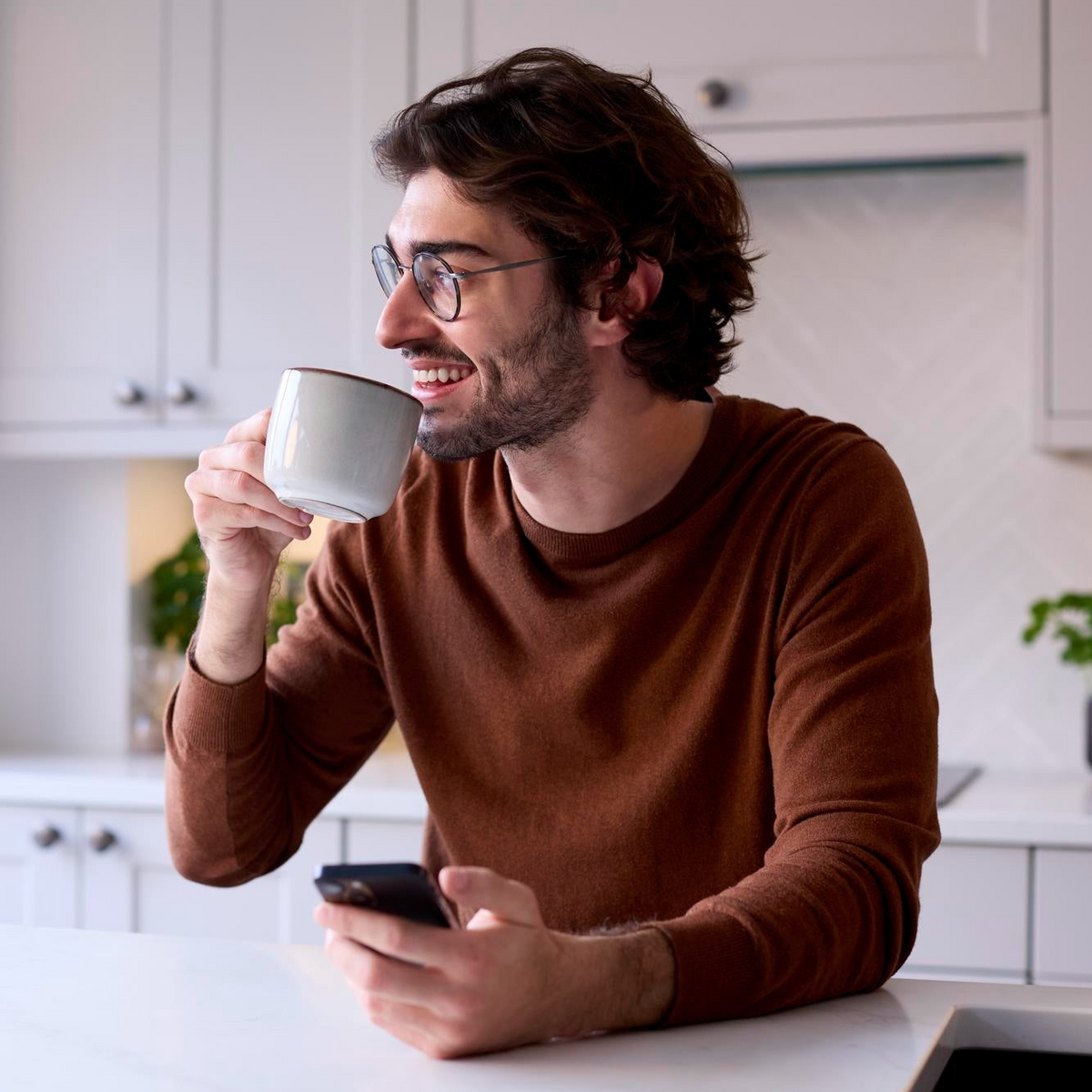 A man with glasses and a beard, wearing a brown long-sleeve shirt, sits at a kitchen counter drinking from a white mug while holding a smartphone.