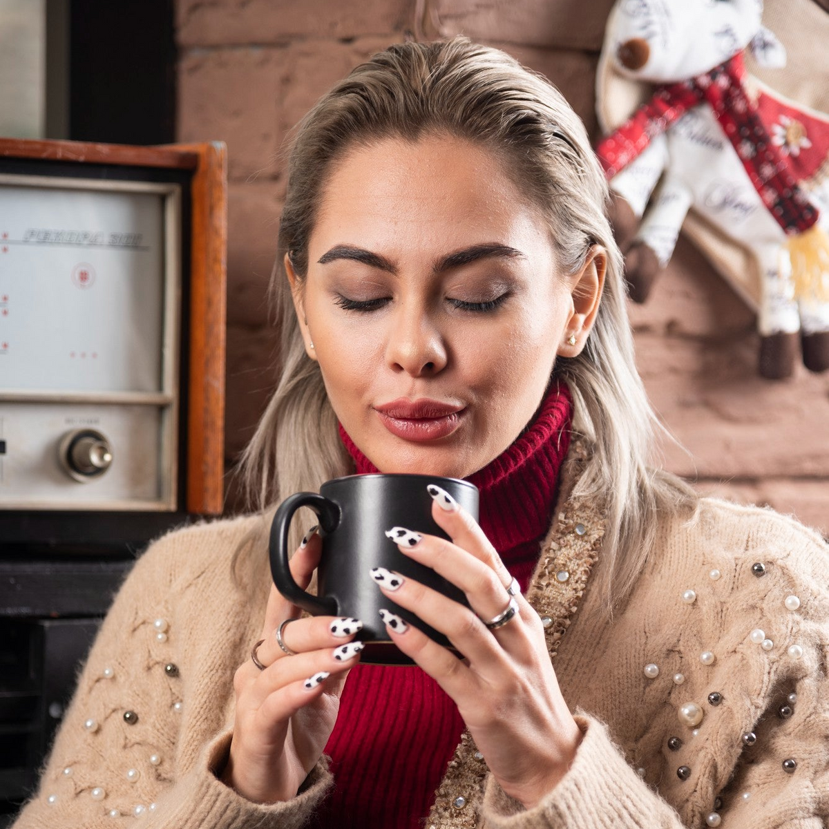 A woman with blonde hair wearing a beige pearl-embellished sweater and a red turtleneck, holding a black ceramic mug with a custom printed design. She is posed in front of a rustic brick wall with a vintage radio and a holiday stocking in the background.