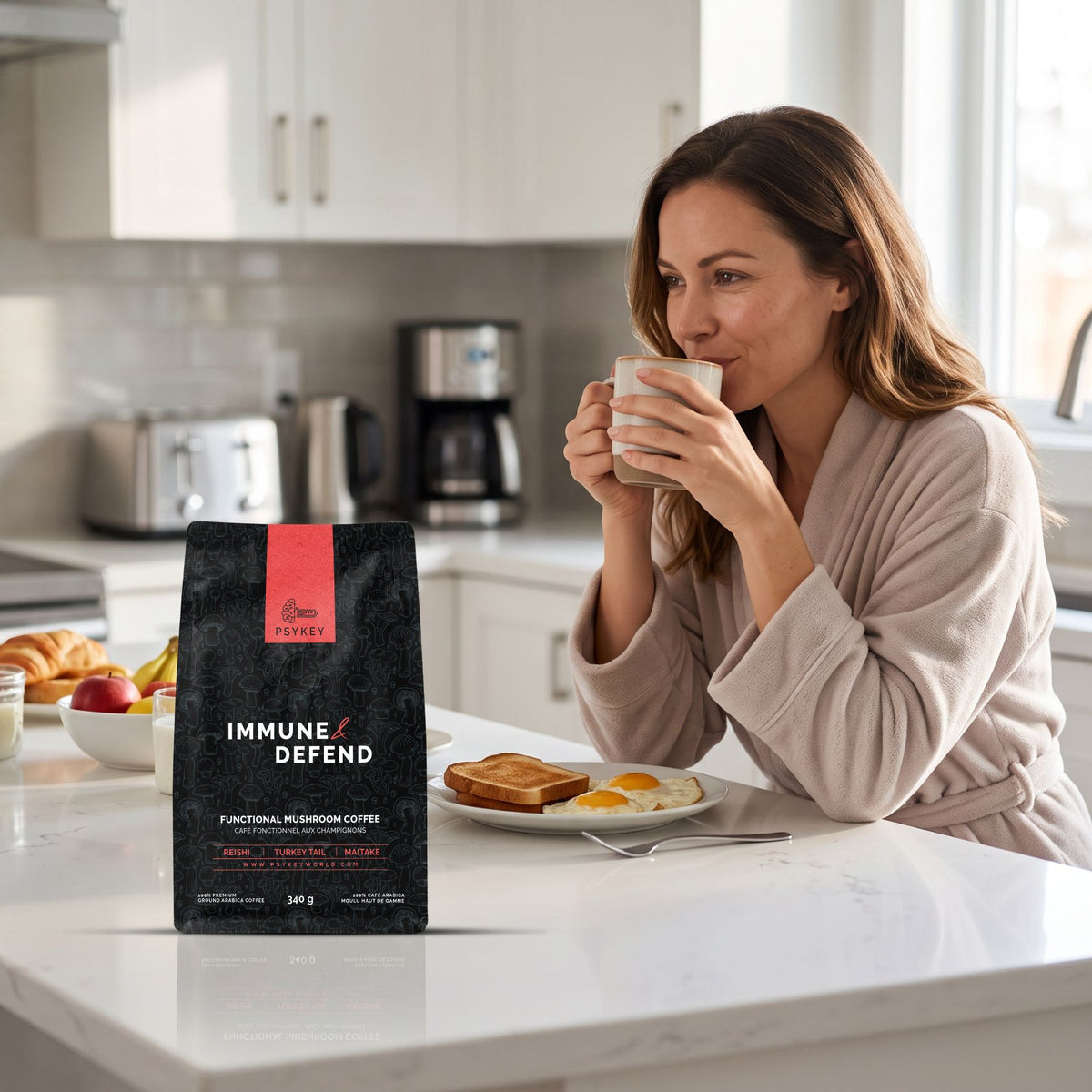 A woman in a cozy bathrobe sitting at a bright kitchen counter drinking coffee next to a bag of PsyKey Immune & Defend mushroom coffee and a plate with toast and eggs.