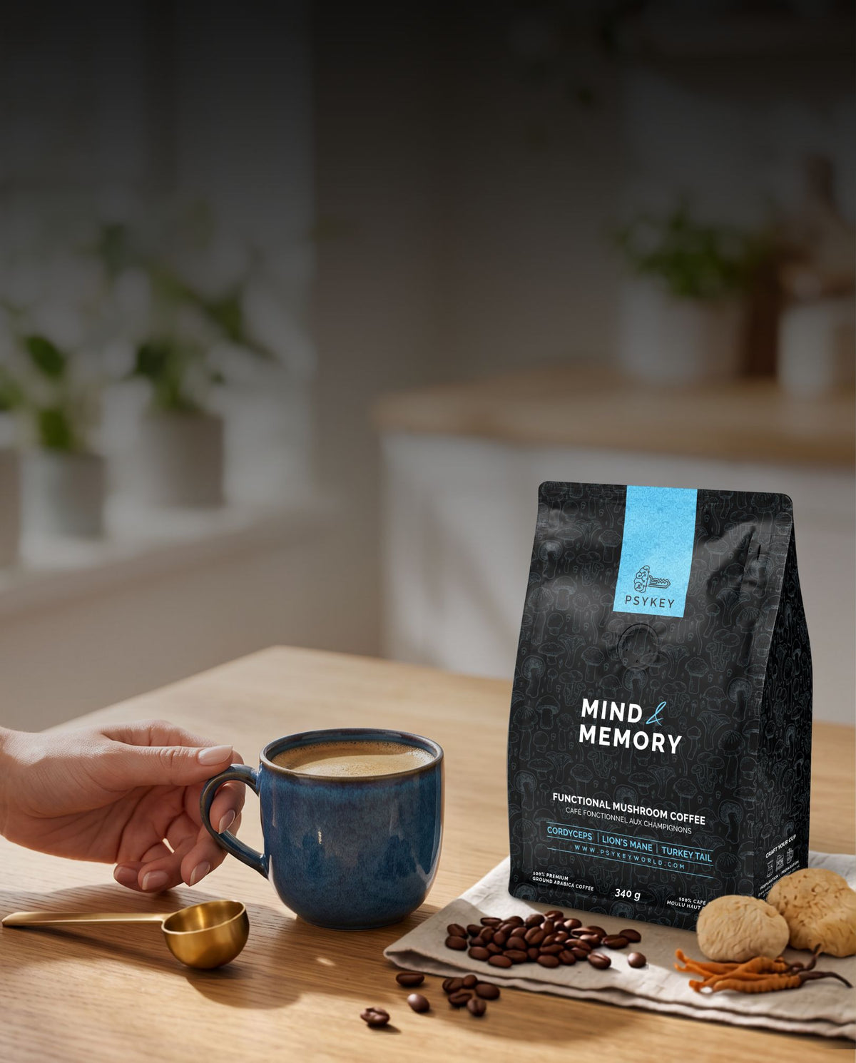 A hand reaching for a blue ceramic mug sitting on a wooden table beside a gold coffee scoop, roasted coffee beans, dried functional mushrooms, and a bag of PsyKey Mind & Memory mushroom coffee.