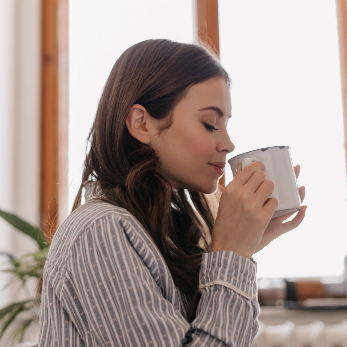 A woman in a striped shirt drinking from a white ceramic mug in a bright indoor setting.
