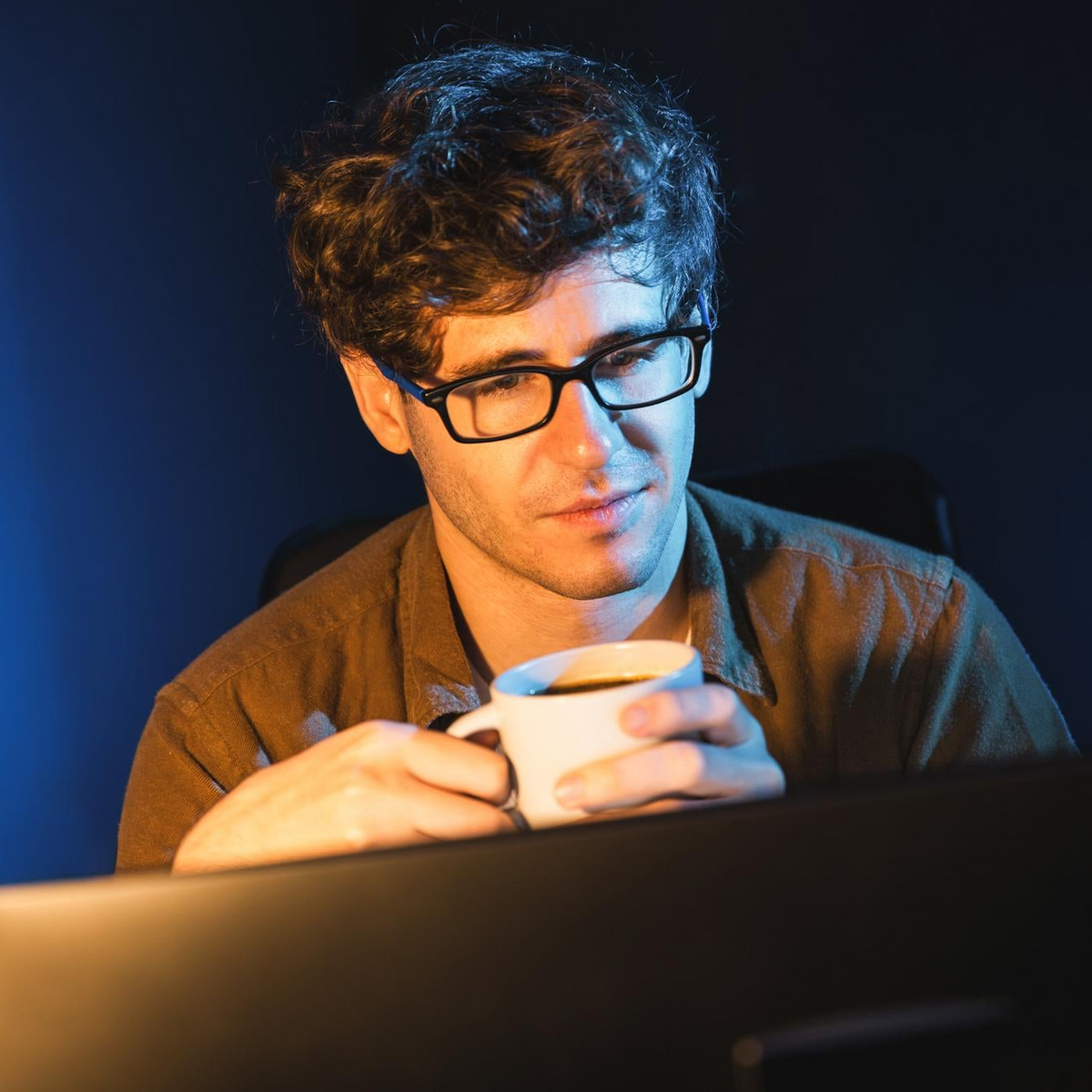 A man with curly dark hair and black-rimmed glasses looks down at a white coffee mug while sitting in front of a computer monitor in a dimly lit room.