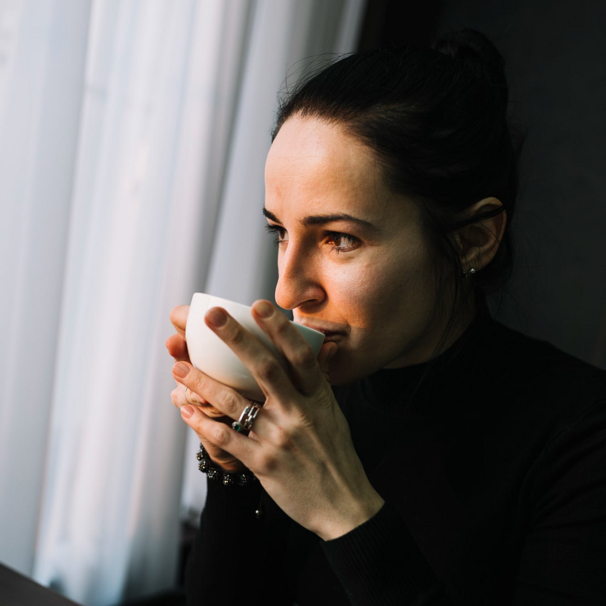 A woman in a black turtleneck sweater holding a white ceramic cup to her lips, captured in soft, warm light against a background of sheer curtains.